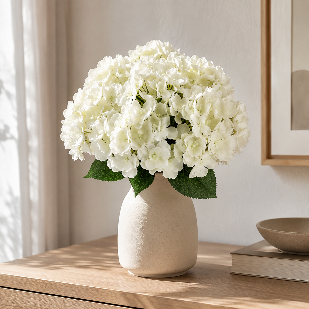 White faux hydrangea flower bouquet in a textured cream ceramic vase, styled on a wooden table in a bright, modern room.