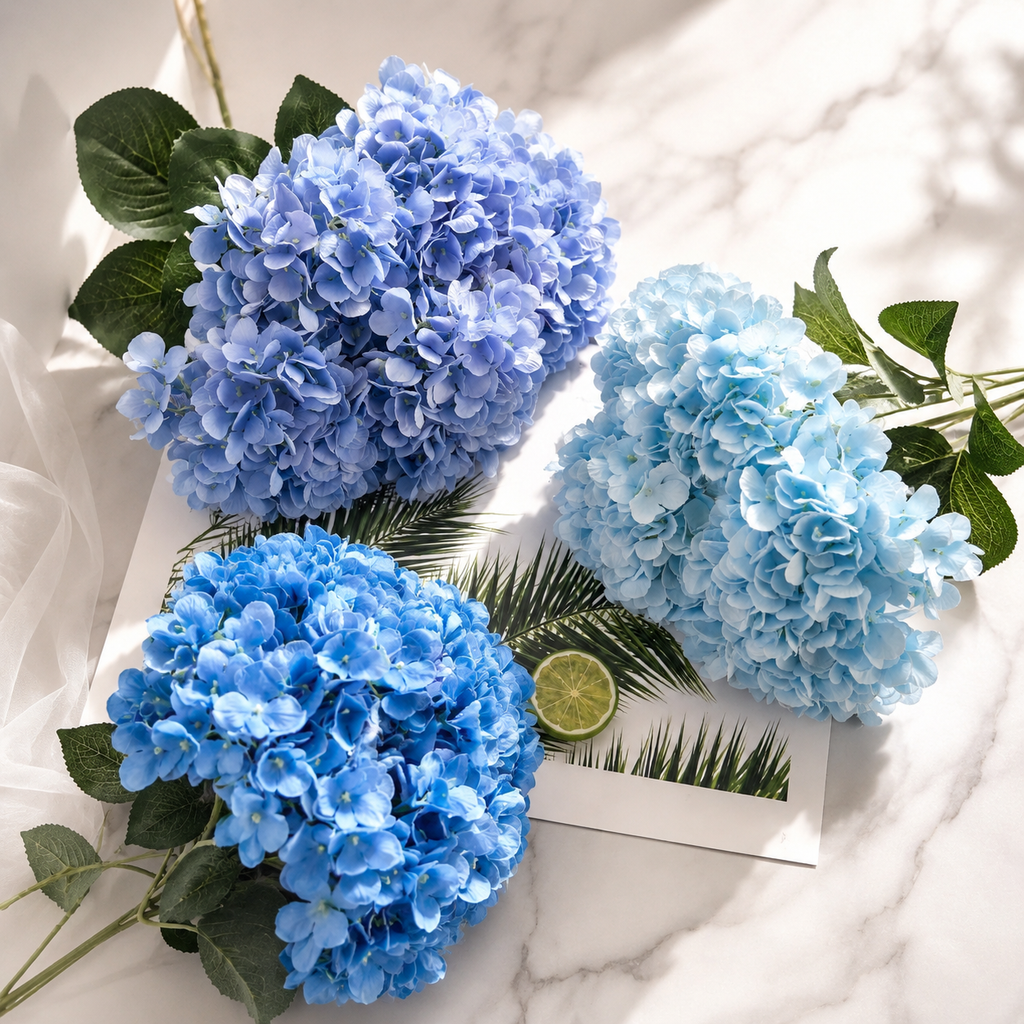 Three artificial hydrangea flower bouquets in shades of blue and light blue, styled on a white marble surface with a small lime slice and a decorative card.